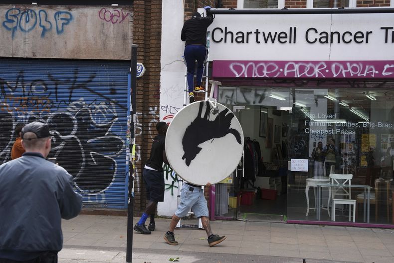 Personas retiran una nueva obra de arte de Banksy, que representa un lobo aullando pintado en una antena parabólica que se colocó en el techo de una tienda en Peckham, al sur de Londres, el jueves 8 de agosto de 2024. (Jordan Pettitt/PA vía AP)