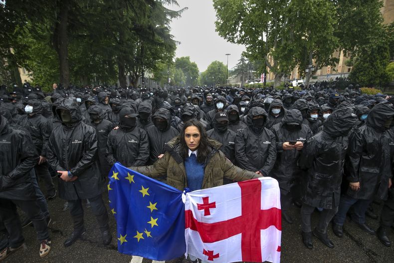 Una mujer sostiene una bandera de Georgia y una de la UE frente a agentes de la policía que impiden el paso a los manifestantes durante una protesta contra una ley cerca del Parlamento en el centro de Tiflis, Georgia, el martes 14 de mayo de 2024. (AP Foto/Zurab Tsertsvadze)