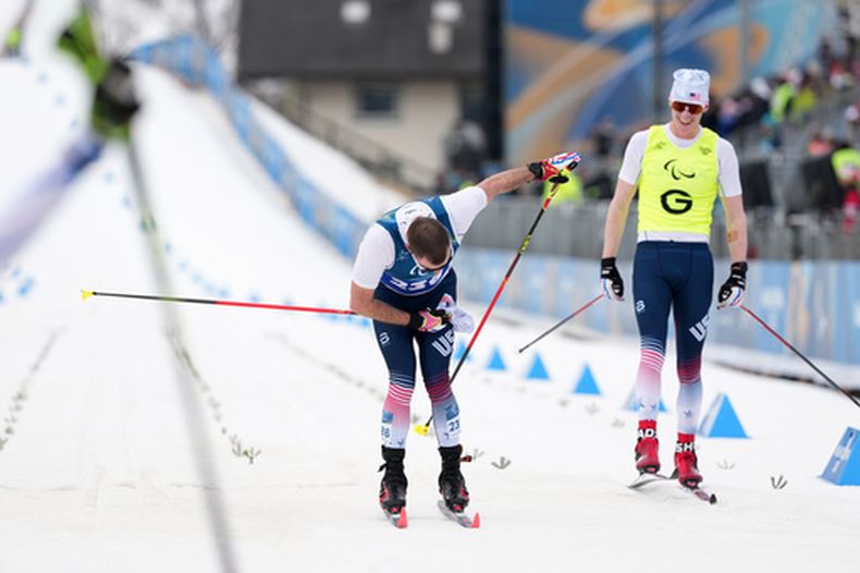 Jake Adicoff cruza la meta, mientras su guía Reid Goble (a la derecha) observa, tras ganar la medalla de oro en la final de esquí de fondo de 10 km con salida a intervalos de la categoría clásica para discapacitados visuales en los Juegos Paralímpicos de Invierno 2026, en Tesero, Italia, el miércoles 11 de marzo de 2026. (Foto AP/Evgeniy Maloletka)