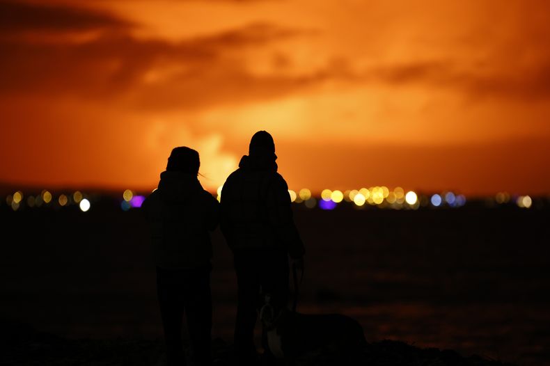 Personas observan el cielo nocturno iluminado por la erupción de un volcán en la península de Reykjanes, en Islandia, el lunes 18 de diciembre de 2023. (AP Foto/Brynjar Gunnarsson)