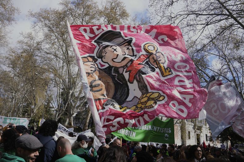 Un grupo de personas marcha con una bandera que muestra a un rico banquero en una protesta contra el alto costo de la vivienda en Madrid, España, el sábado 5 de abril de 2025. (AP Foto/Paul White)