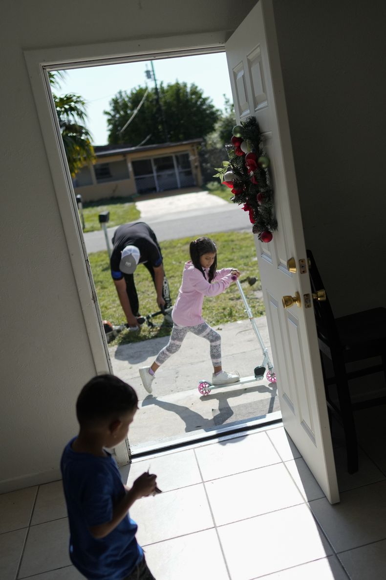Alexa, de 7 años, juega con una patineta que recibió como regalo de Navidad mientras su padre, Alexis Llanos, se prepara para arreglar el jardín, y su hermano Alexis juega dentro de la casa a la que se mudaron en octubre de 2023, cinco años después de haber salido de Venezuela hacia Colombia por amenazas de muerte, el 27 de diciembre de 2023. La de Llanos es una de las primeras familias de migrantes que llegaron a Estados Unidos legalmente bajo un nuevo programa conocido como Oficinas de Movilidad Segura, que busca agilizar el proceso de refugiados. (AP Foto/Rebecca Blackwell)