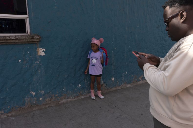 La venezolana Maickeliys Rodríguez, de 6 años, lleva sus nuevas bailarinas plateadas donadas y para un momento a ajustarse la mochila bajo la atenta mirada de un amigo de la familia, haitiano, mientras su familia y otras personas de un refugio migrante acuden a una lavandería cercana en Tijuana, México, el viernes 31 de enero de 2025. (AP Foto/Gregory Bull)