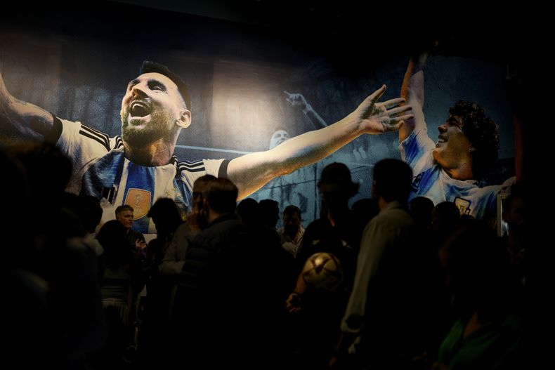 Periodistas pasan frente a las imágenes de Lionel Messi (izquierda) y el fallecido astro Diego Maradona en la exhibición Campeones del Mundo, el miércoles 5 de abril de 2023, en Buenos Aires. La primera exhibición oficial dedicada a los tres títulos mundiales conquistados por Argentina en 1978, 1986 y 2022 ha sido inaugurada en Buenos Aires. (AP Foto/Natacha Pisarenko)