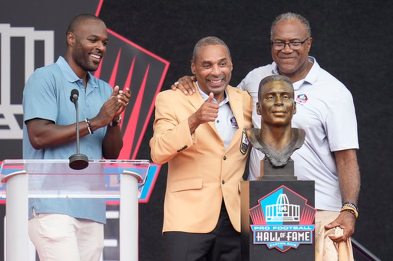 El exjugador de la NFL Roger Craig, al centro, devela su busto junto a su hijo, Roderick Craig, a la izquierda, y Jamie Williams durante la ceremonia de inducción al Salón de la Fama del Fútbol Americano Profesional en el Salón de la Fama del Fútbol Americano Profesional, en Canton, Ohio, el sábado 8 de agosto de 2026. (Foto AP/Sue Ogrocki)