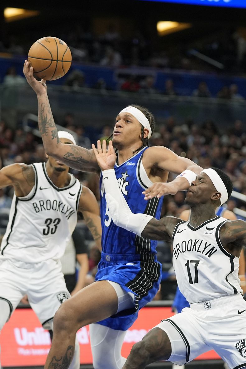 Paolo Banchero, centro, del Magic de Orlando, ataca la canasta pasando al lado de Nic Claxton (33) y Dennis Schroder (17), de los Nets de Brooklyn durante la segunda mitad del juego de baloncesto de la NBA, el miércoles 13 de marzo de 2024, en Orlando, Florida. (AP Foto/John Raoux)