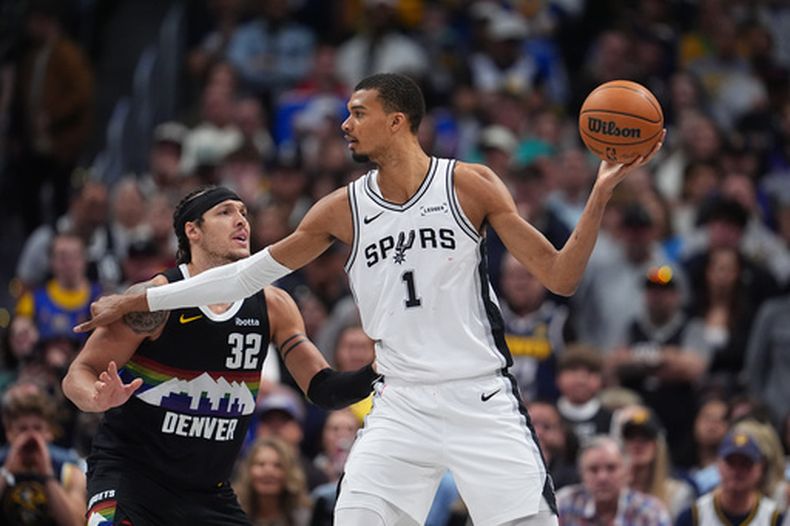 Victor Wembanyama (derecha), centro de los Spurs de San Antonio, busca pasar el balón mientras Aaron Gordon, delantero de los Nuggets de Denver, defiende durante el tiempo extra de un juego de baloncesto de la NBA, el sábado 4 de abril de 2026, en Denver. (Foto AP/David Zalubowski)