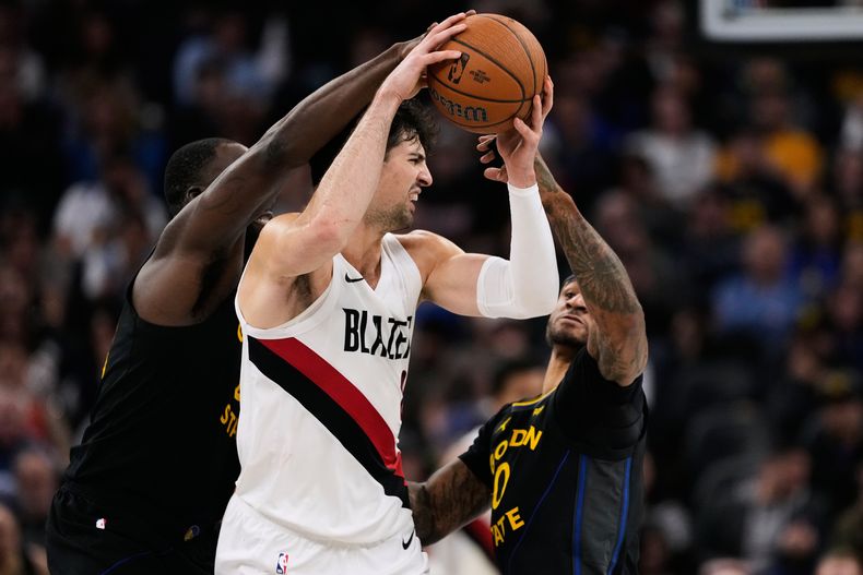 Deni Avdija, de los Trail Blazers de Portland, trata de conservar el balón frente a Draymond Green (izquierda) y Gary Payton II, de los Warriors de Golden State, en el partido del viernes 21 de noviembre de 2025 (AP Foto/Godofredo A. Vásquez)