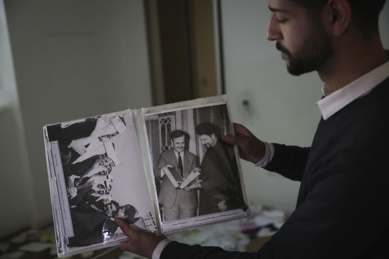 Un hombre muestra viejas fotos del difunto presidente sirio Hafez Assad mientras la gente busca pertenencias en la residencia privada saqueada del presidente sirio Bashar Assad en el distrito de Malkeh de Damasco, Siria, el domingo 8 de diciembre de 2024. (AP Foto/Ghaith Alsayed)