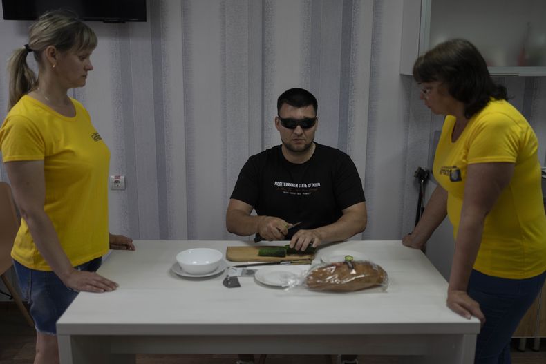 Flanqueado por dos entrenadoras, Denys Abdulin, un exsoldado ucraniano que quedó ciego en la guerra, aprende a usar un cuchillo durante una clase de cocina en un centro de rehabilitación para soldados que perdieron la vista en el campo de batalla, el viernes 21 de julio de 2023, cerca de Rivne, Ucrania. (AP Foto/Jae C. Hong)