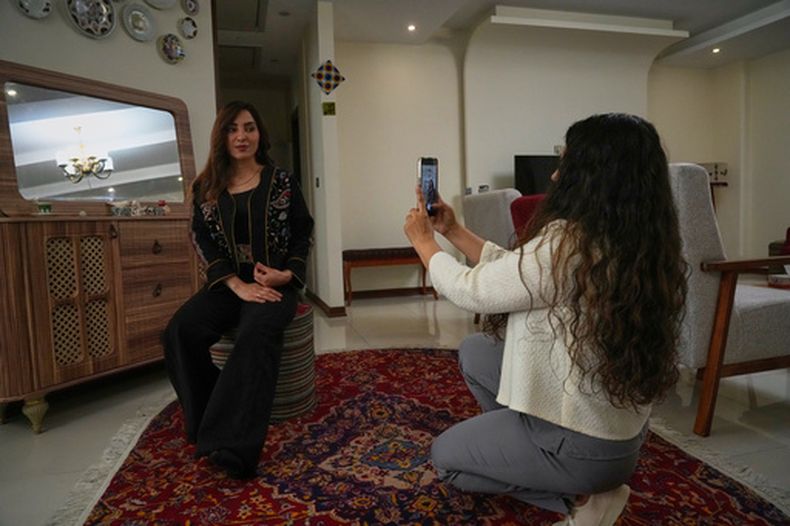 La diseñadora de moda Amen Khademi toma una foto con su celular a la modelo Farnaz Ojaghloo, que lleva una chaqueta diseñada por ella con motivos de inspiración persa, en su estudio en Teherán, Irán, el 23 de abril de 2026. (AP Foto/Vahid Salemi)