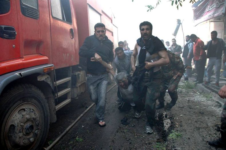 En esta imagen proporcionada por la agencia oficial de noticias siria SANA, soldados gubernamentales, derecha, y un civil, izquierda, trasladan hacia una ambulancia a un hombre herido por la explosi&oacute;n de un carro-bomba en una calle comercial en Hom