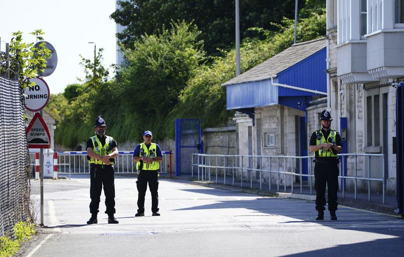 Policías a la entrada del puerto de Portland a donde arribará un autobús del gobierno con solicitantes que serán trasladados a una barcaza de alojamiento, en Dorset, Inglaterra, el lunes 7 de agosto de 2023. (Ben Birchall/PA vía AP)