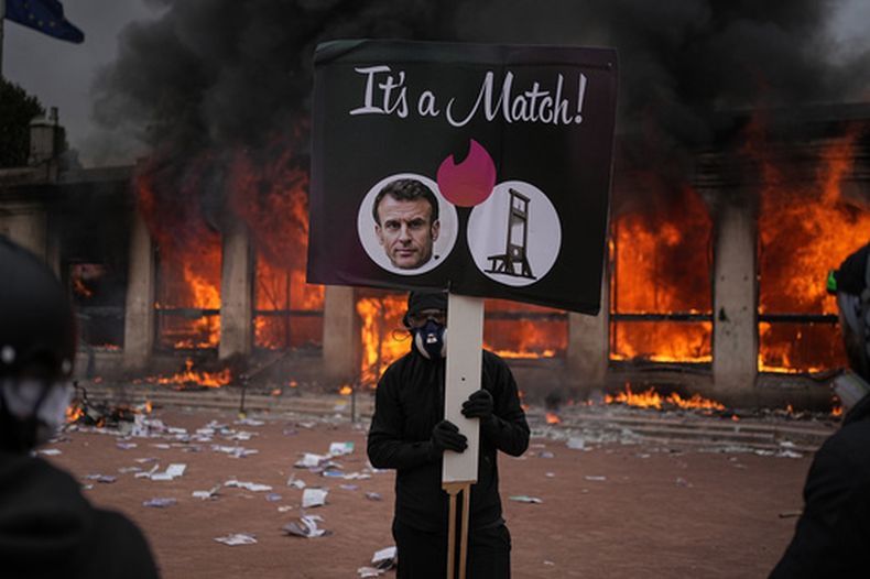 Un manifestante sostiene un cartel con una foto del presidente de Francia, Emmanuel Macron, y una guillotina delante de una oficina de turismo en llamas durante una manifestación contra los costes de la vida y en solidaridad con reclamaciones estudiantiles en Lyon, Francia, el sábado 17 de octubre 2026. (AP Foto/Laurent Cipriani)