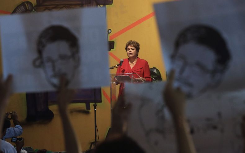 Manifestantes sostienen en lo alto imagenes de Edward Snowden al momento en que irrumpen en una ceremonia en la que la presidenta Dilma Rousseff, al fondo, pronunciaba un discurso de fin de a&ntilde;o ante trabajadores y personas sin casa en Sao Paulo, Br