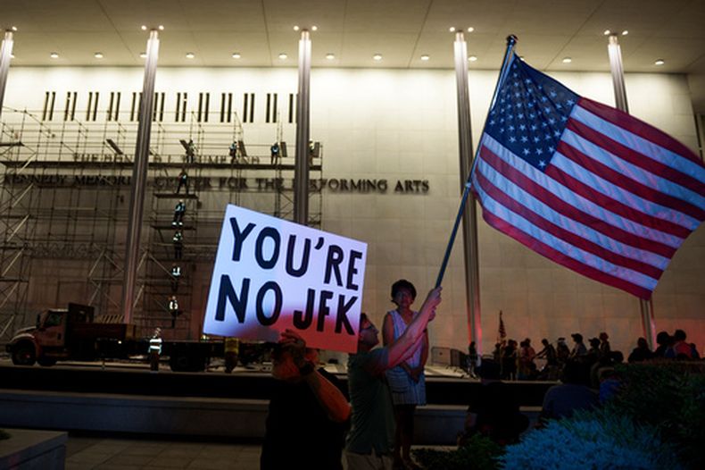 Manifestantes ondean una bandera de Estados Unidos y carteles mientras operarios se preparan para retirar el nombre del presidente Donald Trump del John F. Kennedy Center for the Performing Arts en Washington, el 13 de junio de 2026. (AP Foto/Cliff Owen)