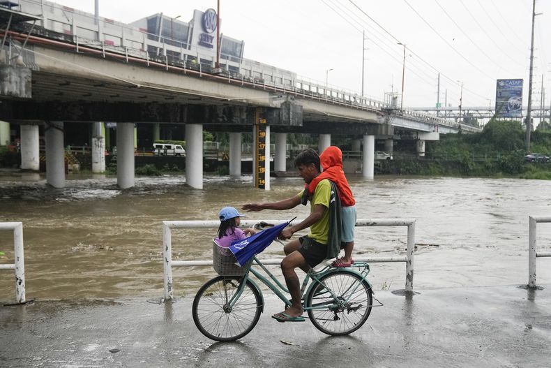 Un hombre y dos niños montan en bicicleta junto a un río crecido por las lluvias del tifón Doksuri el jueves 27 de julio de 2023 en la ciudad de Marikina, Filipinas. (AP Foto/Aaron Favila)