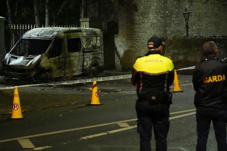 Agentes de la Garda Siochana hacen guardia ante un vehículo policial calcinado cerca del hotel Citywest, el hotel de Dublín que alberga a solicitantes de asilo y en cuyo exterior se registraron disturbios, el 22 de octubre de 2025. (Niall Carson/PA vía AP)