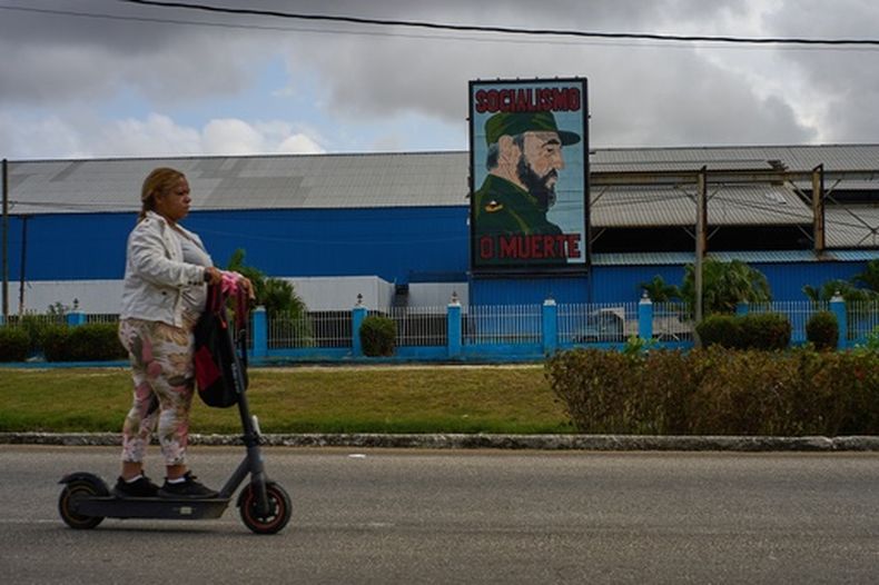 Una mujer pasa en patineta eléctrica frente a una fábrica que exhibe una imagen del fallecido líder cubano Fidel Castro, con la leyenda Socialismo o muerte, en La Habana, Cuba, el jueves 19 de marzo de 2026. (Foto AP/Ramón Espinosa)