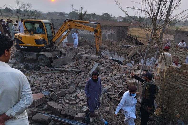 Un trabajador despeja escombros con una excavadora mientras un policía y vecinos de la zona se reúnen en el lugar de un ataque suicida contra un puesto de seguridad en Fatah Khel, en Bannu, un distrito en la provincia paquistnaí de Khyber Pakhtunkhwa, en la frontera con Afganistán, el domingo 10 de mayo de 2026. (AP Foto/Aamad Khattak)