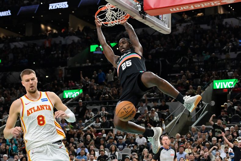 El dominicano David Jones García, de los Spurs de San Antonio, cuelga del aro tras realizar una volcada frente a Kristaps Porzingis, de los Hawks de Atlanta, el jueves 20 de noviembre de 2025 (AP Foto/Darren Abate)