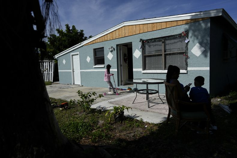 Alexa Llanos, de 7 años, juega con una patineta que recibió de regalo de Navidad mientas su madre, Diomaris Barboza, y su hermano Alexis permanecen sentados bajo la sombra afuera de la casa de Lehigh Acres, en Florida, a la que se mudaron en octubre de 2023, cinco años después de haber salido de Venezuela hacia Colombia por amenazas de muerte, el 27 de diciembre de 2023. La de Llanos es una de las primeras familias de migrantes que llegaron a Estados Unidos legalmente bajo un nuevo programa conocido como Oficinas de Movilidad Segura, que busca agilizar el proceso de refugiados. (AP Foto/Rebecca Blackwell)