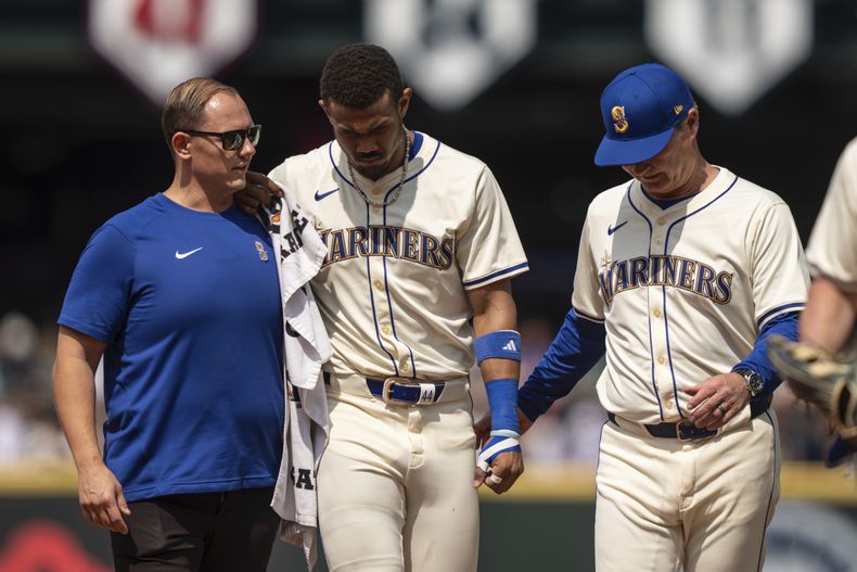 El jardinero central dominicano de los Marineros de Seattle, Julio Rodríguez (centro) es ayudado a salir del campo por el mánager Scott Servais (derecha), y por un integrante del cuerpo técnico durante la sexta entrada del juego de béisbol ante los Astros de Houston, el domingo 21 de julio de 2024, en Seattle. (AP Foto/Stephen Brashear)