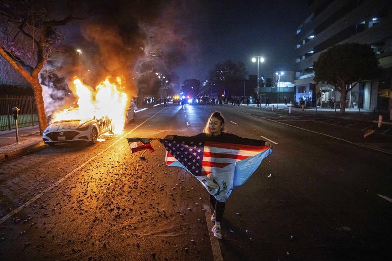 Una persona con varias banderas camina junto a un coche en llamas durante las protestas por las redadas de inmigración de la administración Trump en Los Ángeles, el lunes 9 de junio de 2025. (Foto AP/Ethan Swope)
