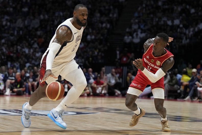 LeBron James, izquierda, de Estados Unidos, y Dennis Schroeder, de Alemania, disputan un balón durante un partido de preparación de baloncesto entre Estados Unidos y Alemania, en la Arena O2 en Londres, el lunes 22 de julio de 2024. (AP Foto/Alastair Grant)