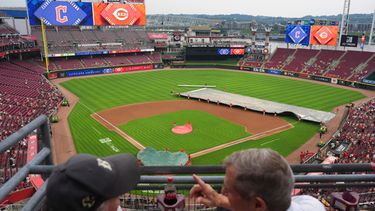 Fanáticos platican mientas que una cuadrilla de trabajadores de los Rojos de Cincinnati cubren el diamante durante el retraso por lluvia previo al partido contra los Guardianes de Cleveland, el lunes 27 de julio de 2026, en Cincinnati. (AP Foto/Kareem Elgazzar)