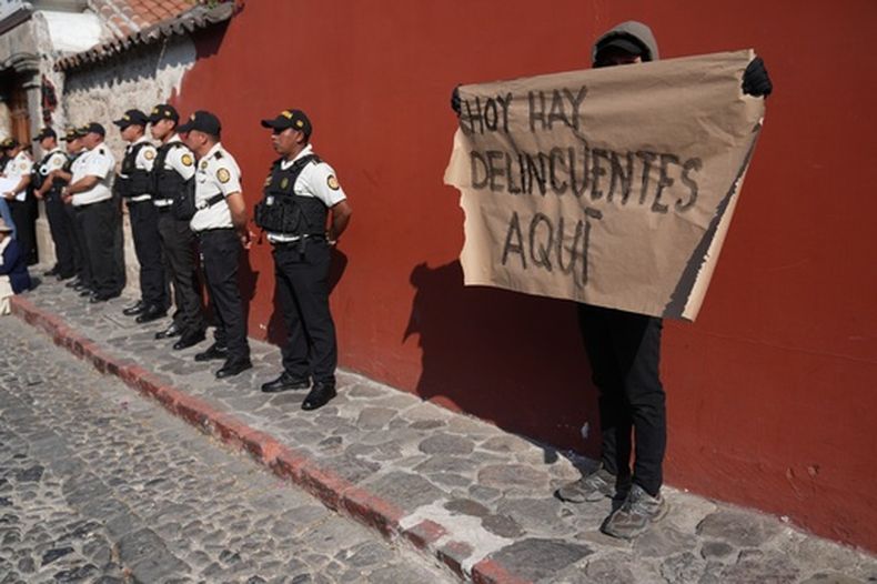 Un manifestante sostiene un cartel junto a la policía que hace guardia en el lugar donde las autoridades de la Universidad de San Carlos se reúnen para nombrar a los jueces de la Corte de Constitucionalidad en Antigua, Guatemala, el lunes 16 de febrero de 2026. (Foto AP/Moisés Castillo)