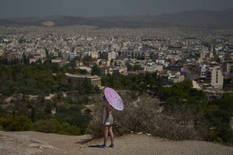 Una turista con sombrilla se ve en la colina de Filopappou con Atenas al fondo, el lunes 7 de julio de 2025, mientras las autoridades en Grecia imponen paros obligatorios en algunos trabajos en lugares donde se esperan más de 40 grados Celsius (104 Fahrenheit). (AP Foto/Petros Giannakouris)