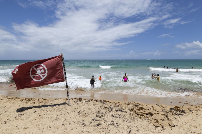 Una bandera de advertencia ondea al viento en una playa mientras la gente se baña en Condado, Puerto Rico, mientras el huracán Erin se acerca, el 15 de agosto de 2025. (AP Foto/Alejandro Granadillo)