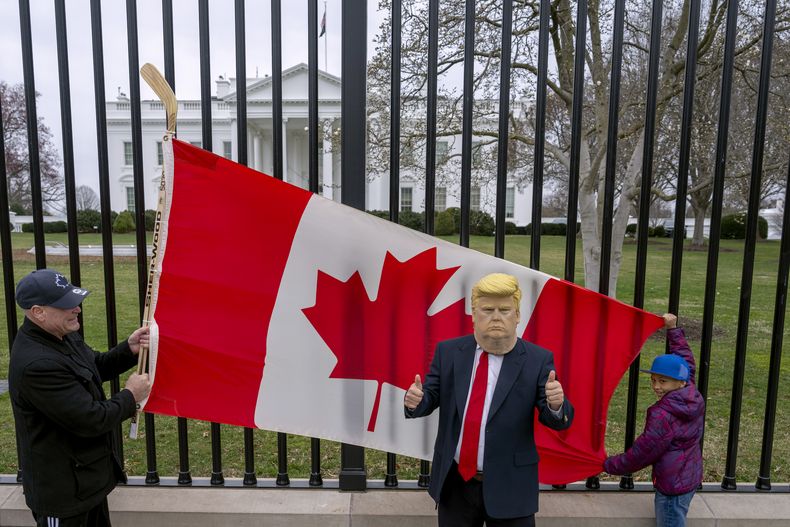 Los residentes de Toronto, Douglas Bloomfield (izquierda) y su hijo Phoenix (derecha), quienes están de vacaciones en Washington, sostienen una bandera canadiense y un palo de hockey para mostrar su apoyo a Canadá mientras otra persona que también se encuentra visitando la ciudad porta una máscara del presidente Donald Trump, frente a la Casa Blanca, el jueves 13 de marzo de 2025. (AP Foto/Ben Curtis)