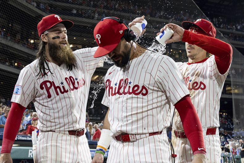 Kyle Schwarber, centro, bateador designado de los Filis de Filadelfia, recibe un baño con vasos de agua por parte de Brandon Marsh, izquierda, y Bryson Stott después de la victoria de su equipo sobre los Marlins de Miami por 9-5, el miércoles 14 de agosto de 2024, en Filadelfia. (AP Foto/Laurence Kesterson)