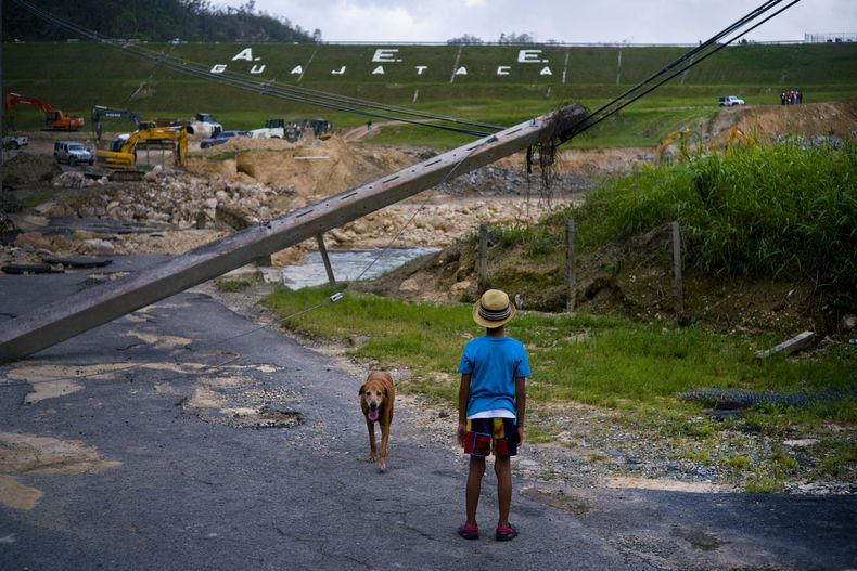 ARCHIVO - Un niño acompañado por su perro observa el martes 17 de octubre de 2017 las labores de reparación de la represa Guajataca, que fue dañada por el huracán María, en Quebradillas, Puerto Rico. (AP Foto/Ramon Espinosa, Archivo)