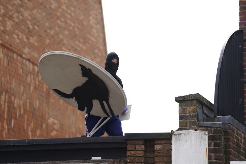 Una persona con el rostro cubierto quita una nueva obra de arte de Banksy, que representa un lobo aullando pintado en una antena parabólica que se colocó en el techo de una tienda en Peckham, al sur de Londres, el jueves 8 de agosto de 2024. (Jordan Pettitt/PA via AP)
