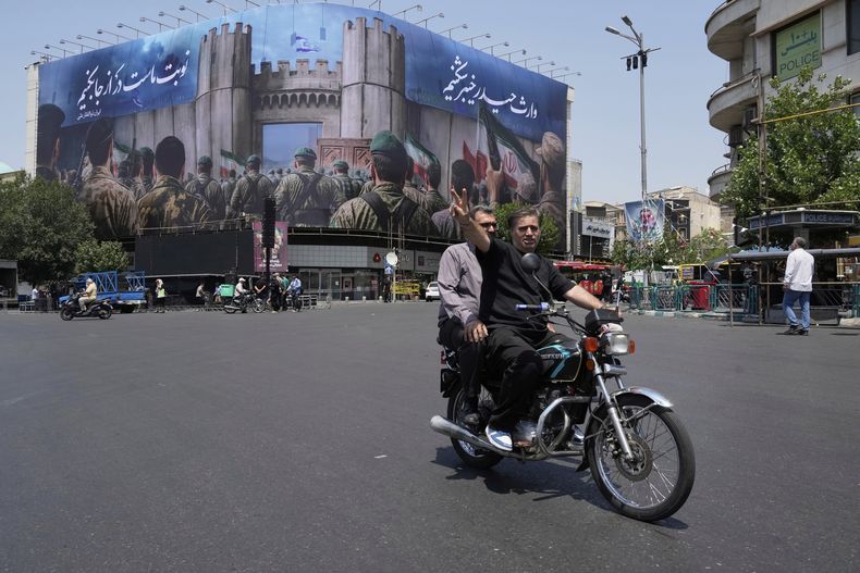 Un hombre hace el signo de la victoria desde su motocicleta tras pasar frente a una manta que muestra a soldados iraníes que se encaminan a atacar territorio israelí, en la Plaza de la Revolución Islámica, el sábado 14 de junio de 2025, en Teherán, Irán. (AP Foto/Vahid Salemi)