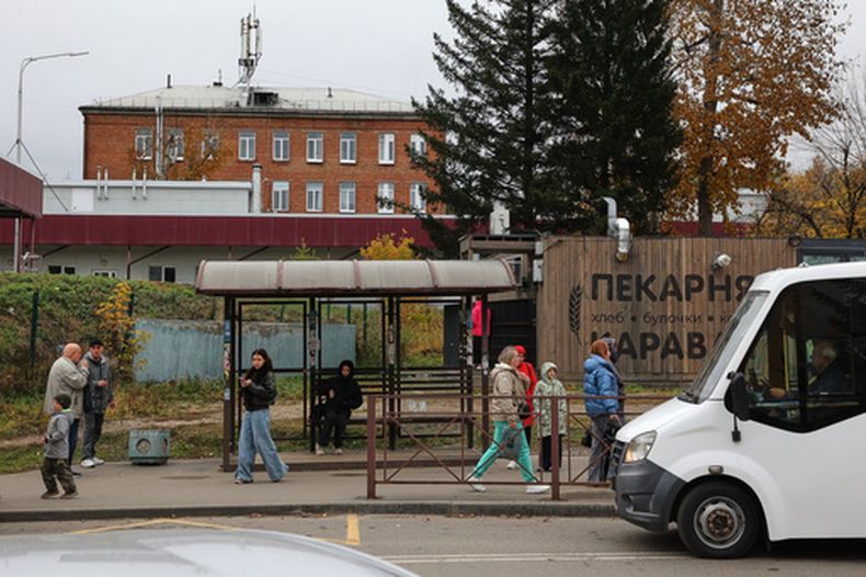 Varias personas en una parada de autobús en Shelekhov, en las afueras de Irkutsk, Rusia, el jueves 8 de octubre de 2026, con el hospital del distrito y el centro médico para el tratamiento de enfermedades infecciosas como fondo. (AP Foto)