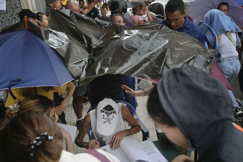 Una mujer, cubierta de la copiosa lluvia por una bolsa de plásticos, busca en una lista los nombres de los votantes para que puedan sufragar durante las elecciones primarias de la oposición en Caracas, Venezuela, el domingo 22 de octubre de 2023. La oposición elegirá un candidato para desafiar al presidente Nicolás Maduro en las elecciones presidenciales de 2024. (Foto AP/Matías Delacroix)