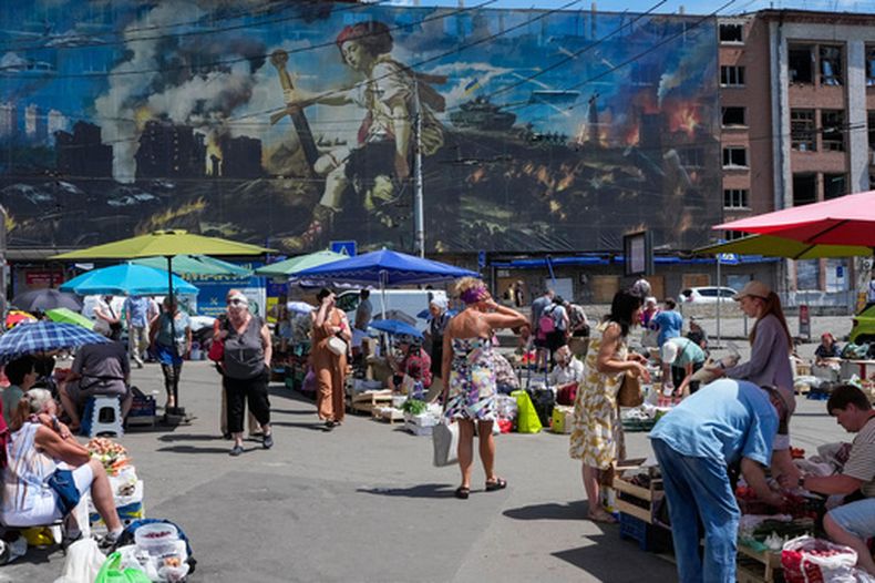 Gente compra comida en un mercado improvisado rodeado de edificios dañados cubiertos por una obra de un artista callejero, junto a un gran mercado destruido hace poco por misiles rusos en Kiev, Ucrania, el lunes 22 de junio de 2026. (AP Foto/Efrem Lukatsky)