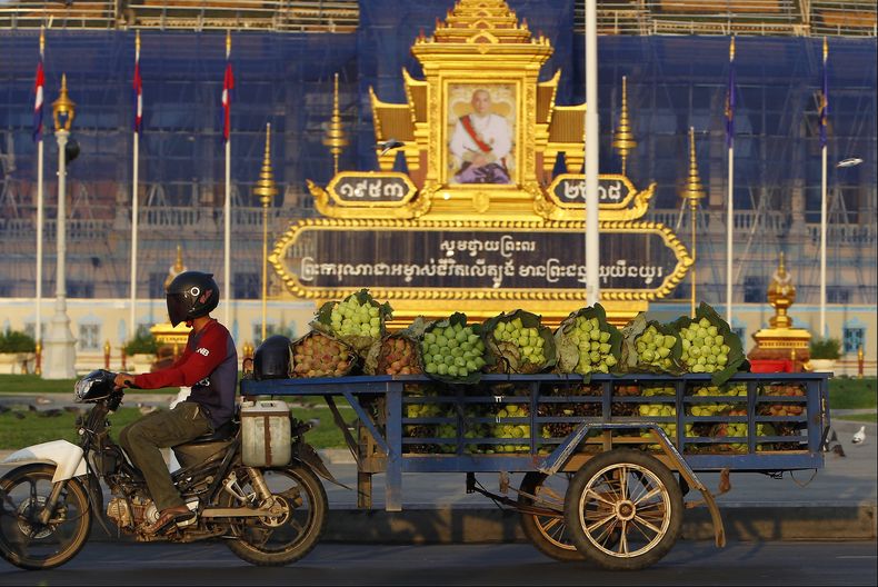 En esta imagen de archivo, un vendedor ambulante que transporta flores de loto pasa por delante del retrato del rey Norodom Sihamoni, frente al Palacio Real, en Nom Pen, Camboya, el 14 de mayo de 2018. (AP Foto/Heng Sinith, archivo)