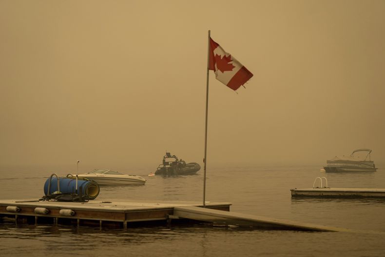 Una densa capa de humo del incendio del lago Lower East Adams llena el aire mientras una bandera canadiense ondea frente a elementos de la Real Policía Montada de Canadá que patrullan en una embarcación en el lago Shuswap, el domingo 20 de agosto de 2023, en Scotch Creek, Columbia Británica, Canadá. (Darryl Dyck/The Canadian Press via AP)