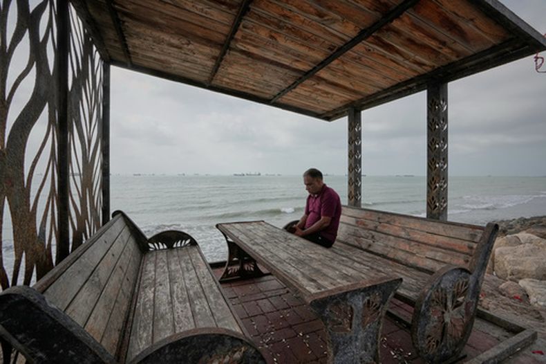 Un hombre descansa en un banco junto a la costa mientras, por detrás, buques comerciales esperan fondeados en el estrecho de Ormuz, frente a la costa de Bandar Abbas, Irán, el 4 de septiembre de 2026. (AP Foto/Vahid Salemi)