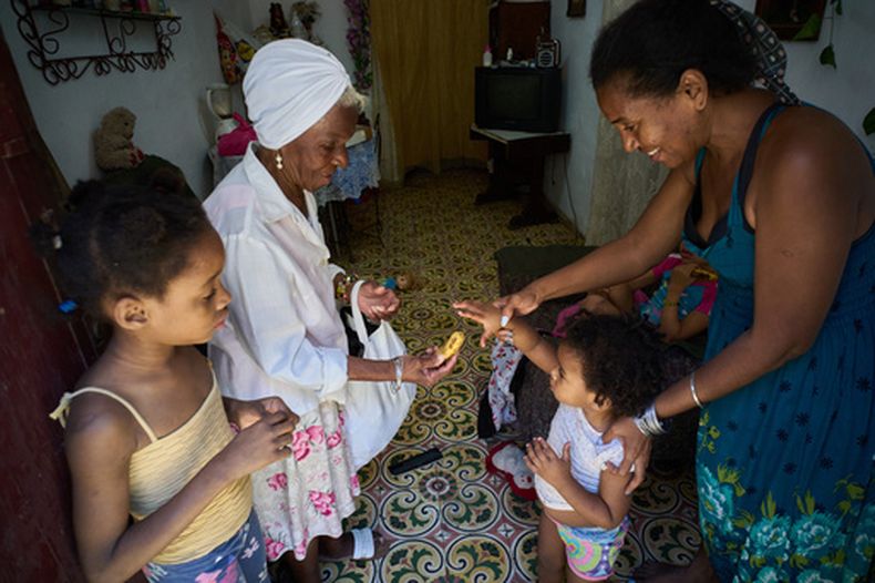 Yuneisy Riviaux ayuda a su hija Seinet a alcanzar un plátano que le ofrece una amiga en su casa de La Habana, Cuba, el miércoles 25 de marzo de 2026. (AP Foto/Ramón Espinosa)