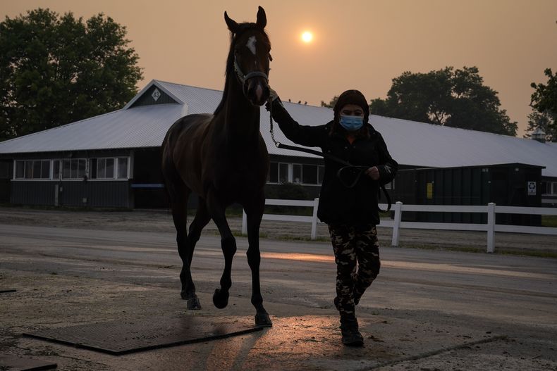 Una cuidadora usa máscara mientras lleva a un caballo de regreso a los establos mientras el sol se oscurece al fondo debido a la neblina causada por los incendios forestales del norte antes de la carrera de Belmont Stakes, el jueves 8 de junio de 2023, en Belmont Park en Elmont, Nueva York. Los entrenamientos y carreras fueron canceladas debido a la mala calidad del aire. (AP Foto/John Minchillo)