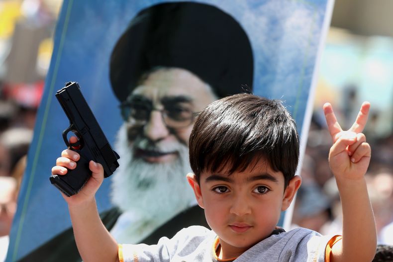 Un ni&ntilde;o iraqu&iacute; que vive en Ir&aacute;n hace la se&ntilde;al de victoria, con una pistola de juguete en la otra mano y una foto del l&iacute;der supremo iran&iacute; ayatola Al&iacute; Jamenei como trasfondo, durante una marcha contra una ofe