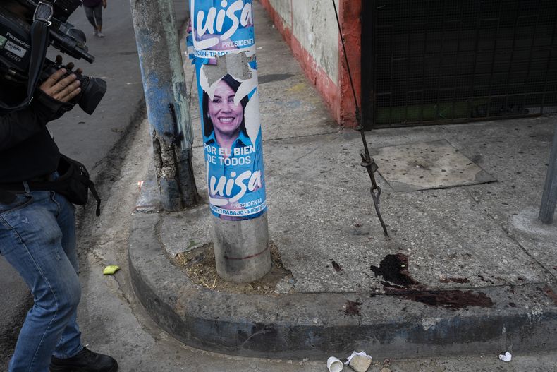 Tras un ataque armado, un reportero de televisión enfoca una mancha de sangre en una vereda, junto a un cartel de la candidata a la presidencia Luisa González, en Guayaquil, Ecuador, el 1 de octubre de 2023. Según los vecinos, dos personas a bordo de una moto dispararon a otras dos, matando a una y causando heridas graves a la otra. (AP Foto/Rodrigo Abd)