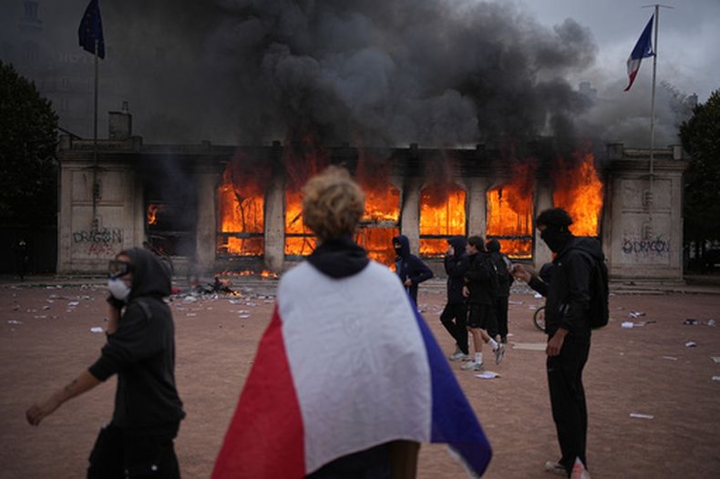 Un manifestante se envuelve con una bandera francesa frente a la oficina de turismo en llamas durante una protesta contra el aumento del costo de vida y en solidaridad con las quejas de los estudiantes en Lyon, Francia, el sábado 17 de octubre de 2026. (Foto AP/Laurent Cipriani)