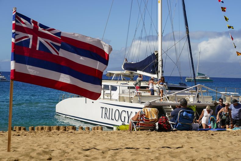 Turistas descienden de un bote de la empresa turística Trilogy Excursions, en la playa Kaanapali, junto a una bandera de Hawai colocada en la arena, el miércoles 6 de diciembre de 2023, en Lahaina, Hawai. (AP Foto/Lindsey Wasson)
